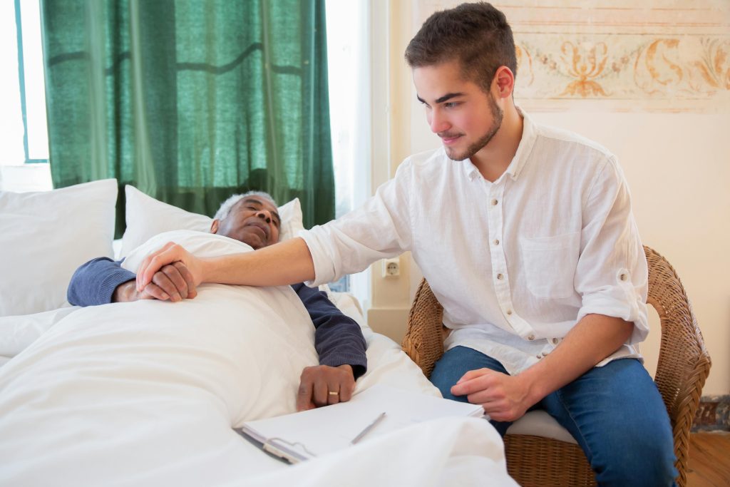 Cuidador le da la mano a adulto mayor acostado en su cama.